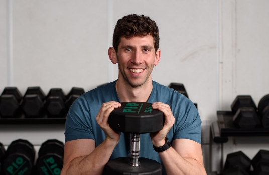 Matthew holding a weight and about to do an exercise, smiling and looking into the camera.